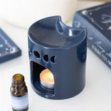 Blue ceramic oil burner with a lit candle inside, next to an essential oil bottle on a white surface.