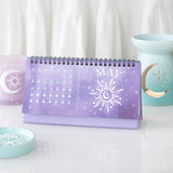 A purple spiral-bound desk calendar featuring a white sun and moon design with the word 'MAY' on the cover, styled on a white surface with decorative pastel props in the background.