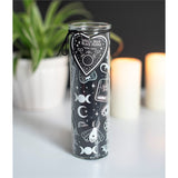 A tall cylindrical glass candle jar with a black label featuring white celestial illustrations, including moon phases, a butterfly, an eye of providence, and a heart-shaped tag that reads 'Vanilla Bean & Black Pepper'. The candle is set on a white surface with a blurred background containing a potted plant and two lit candles.