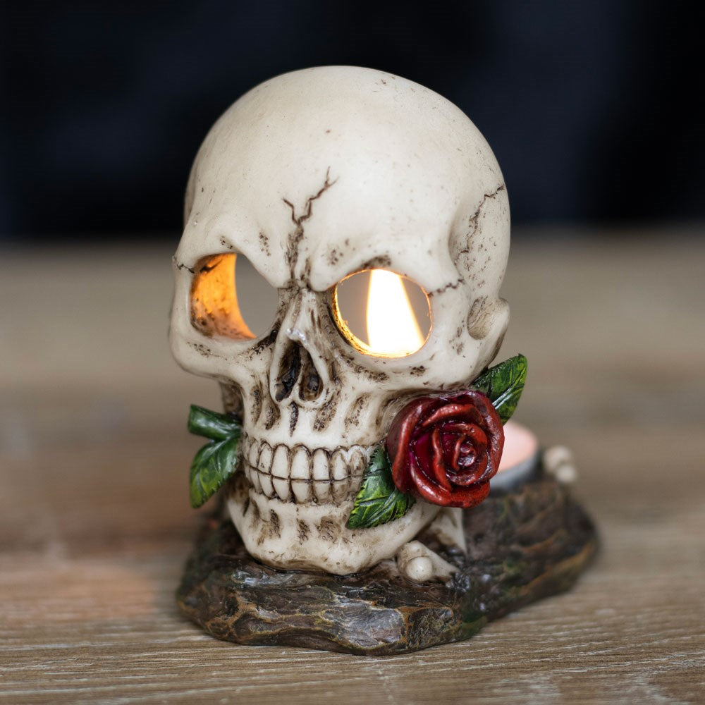 A tealight candle holder in the shape of a skull with a red rose in its mouth and a black base.