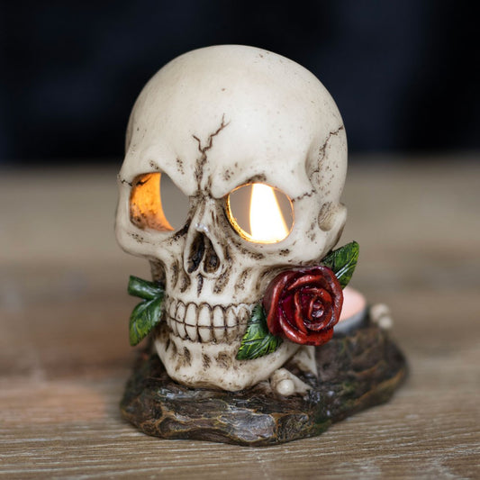 A tealight candle holder in the shape of a skull with a red rose in its mouth and a black base.