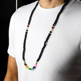 Person wearing a long beaded necklace with black, red, yellow, and multicolor beads on a white shirt.