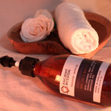 Bottle of Ancient Wisdom massage bath oil with towels and flowers in the background
