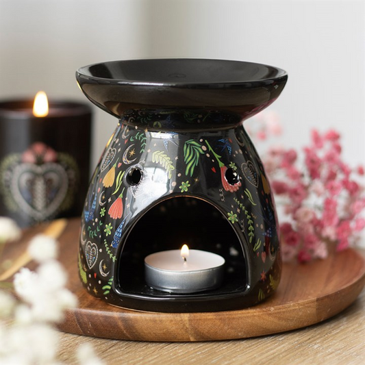Decorative black oil burner with a lit candle on a wooden tray, with a blurred floral background.