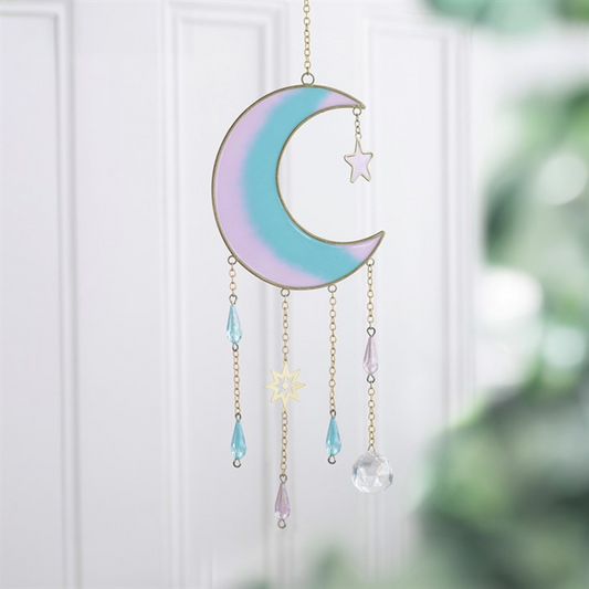 An iridescent pastel crescent moon and star suncatcher hanging in front of a white paneled door. The moon features a soft gradient of lavender, light blue, and mint green with gold-toned metal outlines, while hanging chains dangle light blue teardrop beads, a small gold star, and a clear faceted crystal ball.