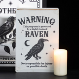 Decorative sign with a raven illustration and humorous text, placed on a surface with a candle and another decorative item in the background.