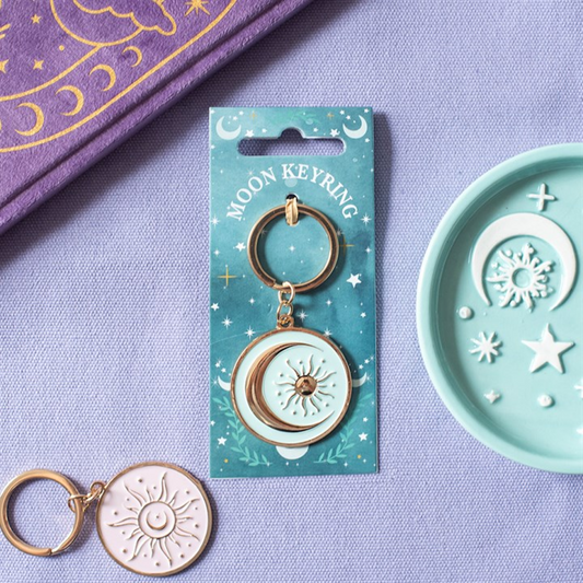 A moon keyring with a gold-tone metal finish and a pale mint moon and sun charm, displayed on its teal backing card. The card is placed on a lavender fabric surface next to a matching mint dish with moon and sun cutouts, and a purple patterned fabric is visible in the top left corner.