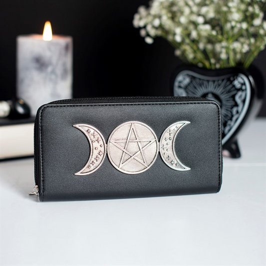 A black zip-around wallet with a silver-toned triple moon and pentagram design on the front, displayed on a white surface with a lit candle and flowers in the background.